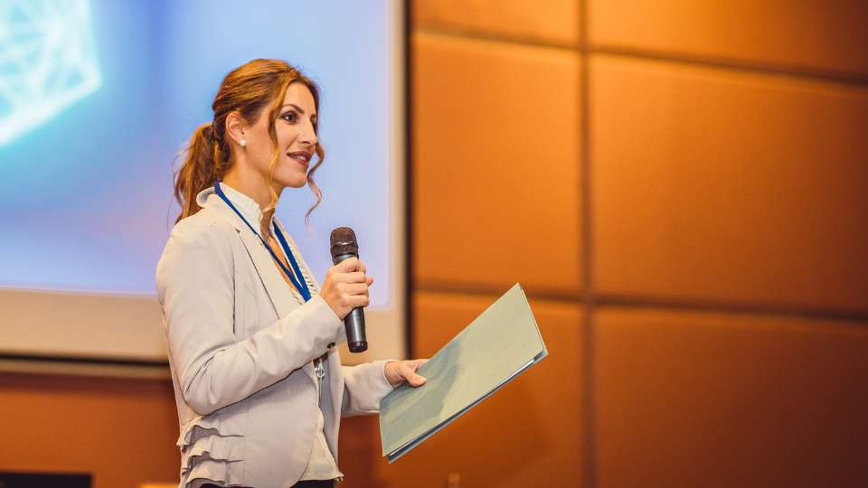 Bild av en moderator som leder ett evenemang med publik och mikrofon.
